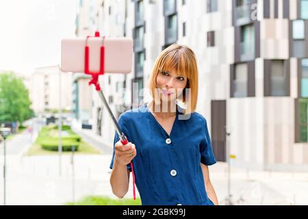 Schlanke, attraktive Frau, die mit ihrem Mobiltelefon auf einem Selfie-Stick ein Selfie auf einer Straße in der Stadt macht, während sie mit einem hohen Schlüssel leise für die Kamera lächelt Stockfoto