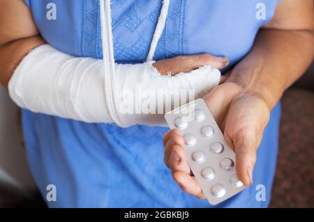 Eine ältere Frau im Ruhestand mit einem zurückgewickelten Arm in einem Guss und einem Verband hält Anäschetabletten. Schlag, Bruch, Knochen, Krankenhaus Stockfoto