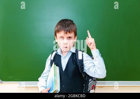 Lustige Schuljunge Grimassen in der Nähe der grünen Schultafel im Klassenzimmer. Grundschulkind mit Tasche und Büchern. Zurück zur Schule. Stockfoto