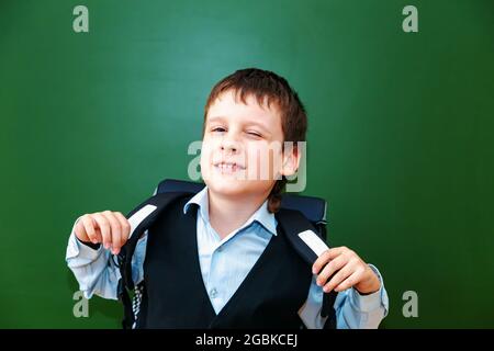 Lustige Schuljunge Grimassen in der Nähe der grünen Schultafel im Klassenzimmer. Grundschulkind mit Tasche. Zurück zur Schule. Stockfoto