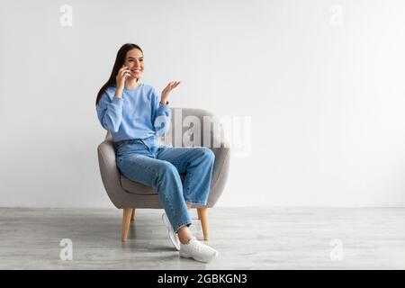 Mobiltelefon-Anruf. Fröhliche junge Frau, die am Handy spricht, im Sessel an der weißen Wand sitzt und Platz für Werbung hat Stockfoto