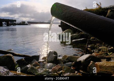 Wasser strömt aus einem rostigen Rohr in den Küstenabschnitt. Sonnenuntergangszeit, am Horizont, Schiffe sind im Hafen. Umweltschutz, Verschmutzung der Welt Stockfoto