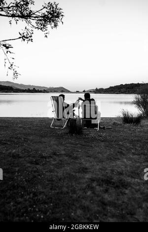 Vertikale Graustufenaufnahme eines Paares auf den Klappstühlen mit dem See im Hintergrund. Stockfoto