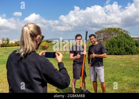 Fitnesstrainer und Klientin posieren für eine Reporterin und schießen auf einem Smartphone. Glücklich und zufrieden. Mit Gymnastikstab. Sommer im Garten. Stockfoto
