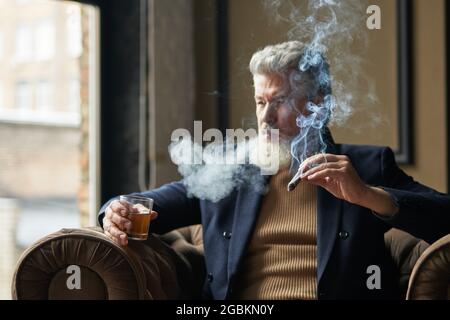 Eleganter, reifer Geschäftsmann, der Zigarre mit Rauch raucht und ein Glas Whisky in der Hand hält, während er sich in einem Stuhl im Loft-Interieur entspannt Stockfoto