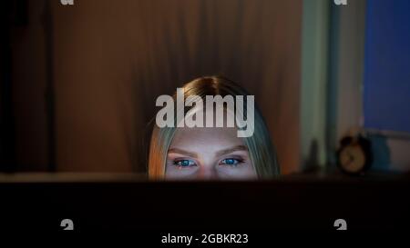 Erntegut Blick auf schöne Frauen sitzen in der Nacht am Computer und mit Blick auf die Kamera über die Oberseite des Monitors mit dunklen Fenster auf unscharfen Hintergrund Stockfoto