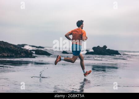Trainieren Sie den Outdoor-Sportler Mann, der auf nassem Sand läuft, am Strand, und trainieren Sie schnell Cardio-Sprints. Profil des Läufers in Sportbekleidung Joggen Stockfoto