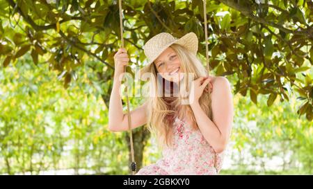 Lächelnde blonde Frau mit Strohhut, schwingt auf der Schaukel im heimischen Garten, Freizeit- und Sommerferienkonzept Stockfoto