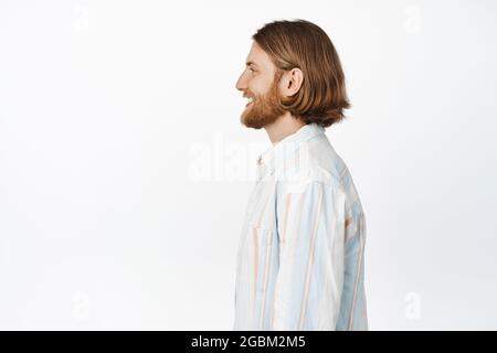 Profilportrait eines erwachsenen, blonden 25er Mannes mit langem Bart, lächelnd, links blickend, im lässigen Hemd vor weißem Hintergrund stehend Stockfoto