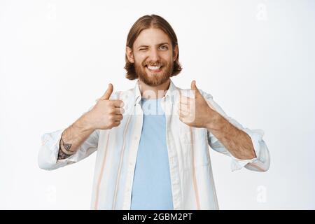 Das Bild des fröhlichen kaukasischen Mannes hat eine positive Meinung, mag und stimmt zu, zwinkend und lächelnd glücklich, zeigt Daumen nach oben in Zustimmung, über Weiß stehend Stockfoto