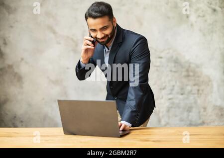 Lächelnder junger indischer Geschäftsmann, der im modernen Büro einen Laptop benutzt, auf dem Smartphone am Arbeitsplatz sitzt und auf den Laptop-Bildschirm schaut, telefoniert der spanische männliche Manager Stockfoto