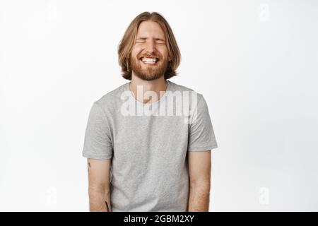 Bild eines sorglosen, gutaussehenden Mannes, blonden Mannes, der lachend und lächelnd mit geschlossenen Augen in einem grauen T-Shirt auf weißem Hintergrund steht Stockfoto