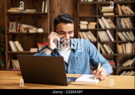 Lächelnder indischer Mitarbeiter, der Laptop für die Fernarbeit verwendet, am Schreibtisch im Heimbüro sitzt, auf dem Smartphone redet und Notizen macht, Eastern man telefoniert, schreibt auf einem Notizblock und benutzt einen Laptop Stockfoto