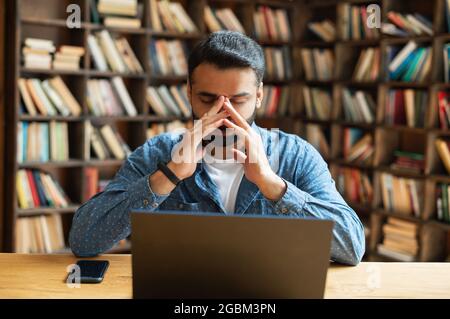 Müde, junger indischer Geschäftsmann, der am Schreibtisch vor dem Laptop sitzt, mit beiden Händen bedeckt sein Gesicht, frustriert ist, müde und depressiv, männliche Angestellte im östlichen Büro, hat Probleme mit dem Projekt Stockfoto