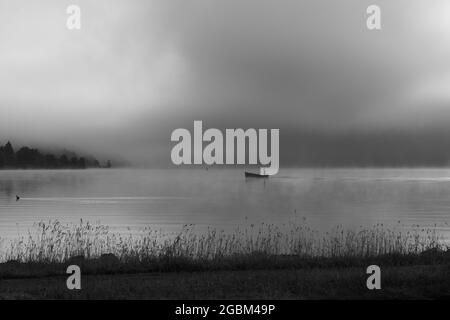 Ein früher Morgen am Lac de Joux, Schweiz: Ein einflüchtiger Fischer auf einem Boot, Nebel versteckt immer noch die aufgehende Sonne Stockfoto