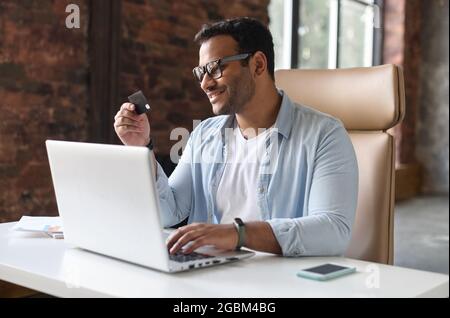Ein indischer Mann sitzt vor dem Laptop und hält eine Kreditkarte, zahlt online, kauft ein, ein Freiberufler aus dem Osten täte Transaktionen, verwendet E-Banking-App auf dem Computer und gibt Daten ein Stockfoto