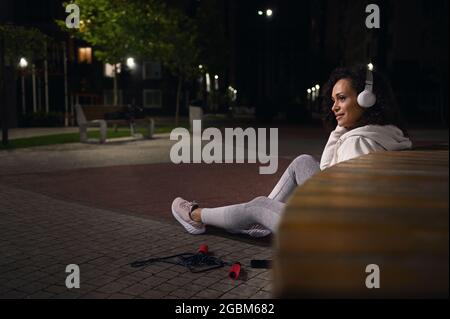 Rückansicht des afroamerikanischen Sportlers mit Kopfhörern, der sich nach dem harten Training am Abend auf einer Holzbank entspannt Stockfoto