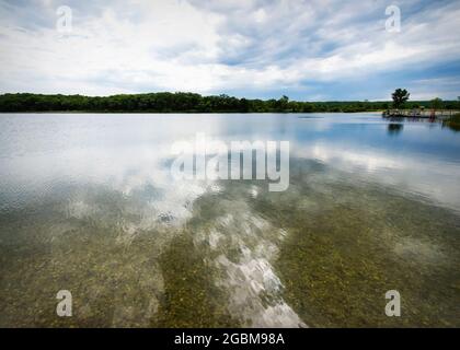 Blick über einen klaren See im Waukesha County, Wisconsin. Angelpier in der Ferne. Wolken spiegeln sich im klaren Wasser. Stockfoto