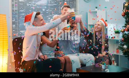 Nahaufnahme der fröhlichen Firma, die Weihnachten im Konfetti-Blasen feiert, während sie zu Hause Jubeleien macht und Partypfeifen bläst. Stockfoto