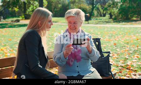 Ältere Großmutter, die ihre Enkelin auf dem Smartphone zeigt. Sie sitzen auf der Bank und reden im Park. Stockfoto