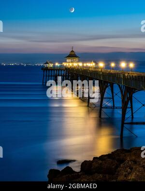 In der Abenddämmerung in Somerset, England, geht ein Halbmond über dem Bristol Channel und dem Clevedon Pier unter. Stockfoto