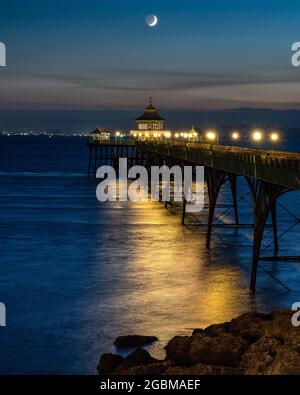 In der Abenddämmerung in Somerset, England, geht ein Halbmond über dem Bristol Channel und dem Clevedon Pier unter. Stockfoto