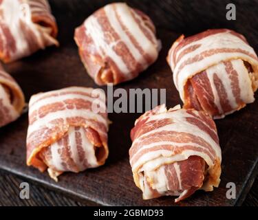 Rohe englische Fleischbällchen, ungekochte Faggots werden aus gehacktem Innereien und Fleisch hergestellt, das in gestreiftem Speck auf einem rustikalen Schneidebrett aus der Nähe eingewickelt ist Stockfoto