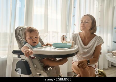 Baby sitzt an einem Kindertisch. Mama füttert das Kind mit Haferbrei. Mutter gibt Babynahrung aus einem Löffel. Stockfoto