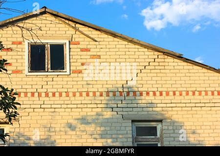 Ein zweistöckiges weißes Ziegelhaus mit einem Riss unter einem Dach. Ein beschädigtes Haus in Privatbesitz gegen blauen Himmel. Verfallene Wände des alten Gebäudes müssen mal wieder aufgesetzt werden Stockfoto