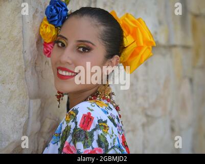 Die junge schöne mexikanische Yucatecan-Volkstänzerin trägt ein traditionelles folkloristisches Kleid mit bunten Blumen im Haar und lächelt für die Kamera. Stockfoto