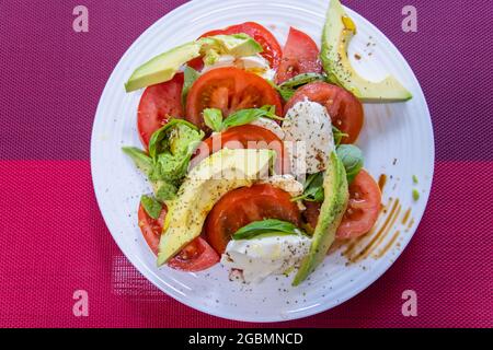 Traditioneller Tricolore-Salat auf einem weißen Teller, Zutaten in Scheiben geschnittene Avocado und Tomaten und weiße Mozzarella mit einem Dressing aus Balsamico-Essig Stockfoto