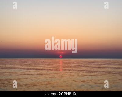 Leuchtend rote Sonne untergeht hinter dem Horizont in einem gradienten Himmel am Meer Stockfoto