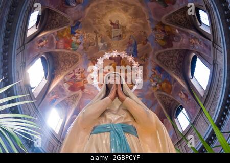 Eine Skulptur, Statue der Jungfrau Maria, mit weißem Gewand, blauer Schärpe und einem beleuchteten Heiligenschein unter einer bemalten Kuppel. In einer lokalen Kirche in Rom, Italien. Stockfoto