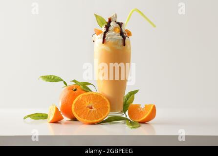 Orangefarbener Milchshake mit cremefarbenem Dekor, verziert mit Früchten und Blättern auf weißem Tisch und hellem, isoliertem Hintergrund. Vorderansicht. Stockfoto