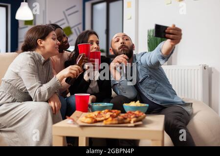 Diverrse Gruppe von Mitarbeitern, die Selfie auf dem Smartphone nach der Arbeit auf der Bürofeier. Fröhliche Kollegen, die Spaß am Feiern haben, indem sie mit Pizza-Chips-Flaschen und Tassen Bier Alkohol trinken Stockfoto