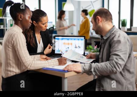 Vielbeschäftigte multikulturelle, vielfältige Mitarbeiter, die jährliche Finanzstatistiken analysieren, sitzen am Schreibtisch vor dem Laptop und halten Dokumente bereit, die nach Geschäftslösungen suchen. Team von Geschäftsleuten, die im Unternehmen arbeiten Stockfoto