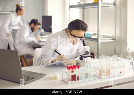 Fokussierte Frau sitzt an einem Tisch im Labor und untersucht die Zellen unter einem Mikroskop. Stockfoto