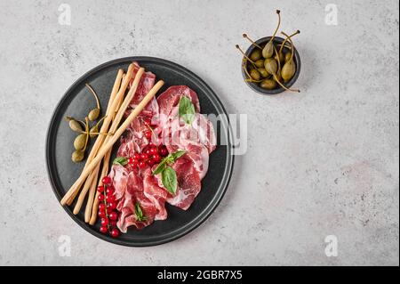 Scheiben Prosciutto coppa di parma Schinken mit Basilikum, Grissini-Sticks, Kapern und roter Johannisbeere auf dunklem Teller auf strukturiertem Hintergrund. Draufsicht mit Kopierbereich Stockfoto