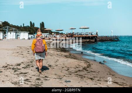 Frau Reisende in einem Sommerhut und Tourist in einem Rucksack Spaziergänge am Strand und genießt eine schöne Aussicht Stockfoto