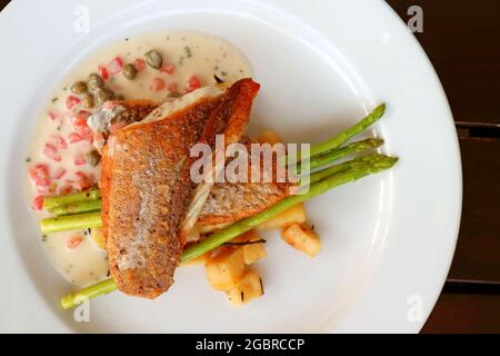 Draufsicht auf den in der Pfanne angebratenen Seebarsch mit Tomatenkaper-Senf-Sahnesauce auf weißem Teller Stockfoto