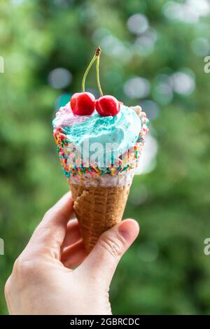 Weibliche Hand hält mehrfarbiger Eiscreme mit Kirschen in einem Waffelkegel vor grünem Hintergrund Stockfoto