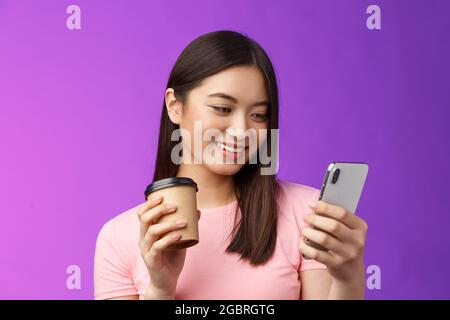 Nahaufnahme zärtliche, freundliche asiatische Bloggerin, die in den sozialen Medien nachschaut, trinkt Kaffee zum Mitnehmen, lächelt fröhlich auf Telefonbildschirm und liest Stockfoto