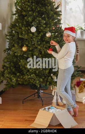 Das Fest steht bevor! Nahaufnahme Seitenansicht Profil Portrait of cute Little girl with long hairstyle with Santa's Hut on Head, excited setting the pinetree, e Stockfoto