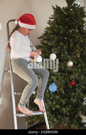 Kleines Mädchen mit Weihnachtsmann Hut sitzt auf der Leiter und schmückt Weihnachtsbaum bunte Kugeln. Neujahrskonzept. Stockfoto