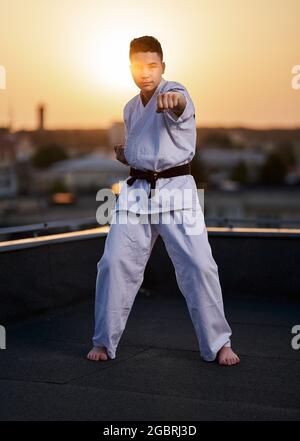 Ein junger junger Karate-Praktizierender im weißen Kimono-Training auf dem Dach bei Sonnenuntergang Stockfoto