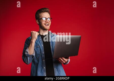 Fröhlich lächelnder junger Mann in legerer Kleidung, der über dem roten Wandhintergrund steht und seinen Laptop hält, um den Erfolg zu feiern Stockfoto