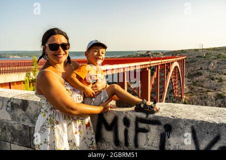 ZADAR, KROATIEN - 10. Jul 2021: Eine Frau, die mit ihrem Kind auf einer langen Brücke über den Fluss Zrmanja in Zadar, Kroatien, an einer Barriere lehnt Stockfoto