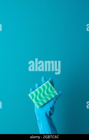 Eine Hand in einem blauen Schutzhandschuh aus Gummi hält einen grünen Waschschwamm auf blauem Hintergrund Stockfoto