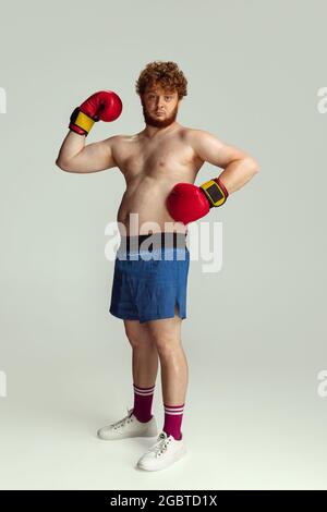 Lustiger rotköpfiger Mann in blauen Boxshorts und Handschuhen isoliert auf grauem Studiohintergrund. Konzept von Sport, Humor und Körper positiv. Stockfoto