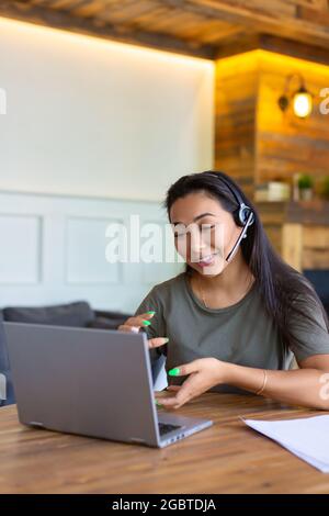 Porträt einer lächelnden asiatischen Frau mit Headset vor dem Laptop-Monitor. Während eines Online-Videoanrufs lächelt und gestikelt sie mit den Händen. Stockfoto